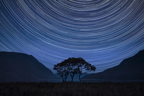 Digital composite image of star trails around Polaris with beautiful foggy misty countryside landscape surrounding crummock water in lake district in england