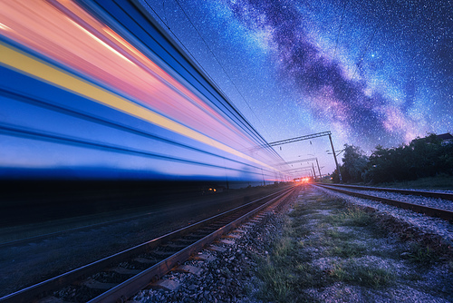 High speed train in motion and Milky Way at starry night. Industrial landscape with sky and stars over blurred modern passenger train and railroad. Railway station and space. Technology and nature