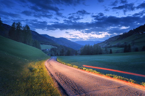 Blurred car headlights on rural road at night in summer. Landscape with asphalt road, light trails, mountains, green grass, trees, and blue sky with clouds at dusk. Roadway through the meadows