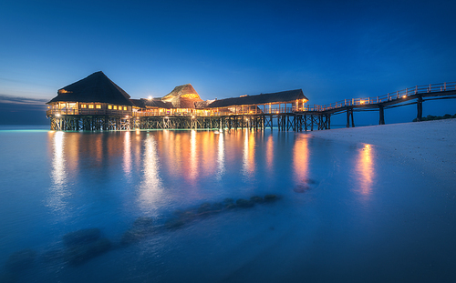 Beautiful wooden restaurant on the water in summer night. Landscape with hotel on the sea, illumination, jetty, sandy beach, blue sky and lights reflected in blurred water at dusk in Zanzibar, Africa