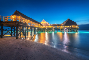 Beautiful wooden restaurant on the water in summer night. Landscape with hotel on the sea, illumination, jetty, sandy beach, blue sky and lights reflected in blurred water at dusk in Zanzibar, Africa