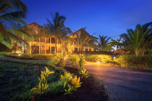 Amazing hotel with lighted paths in flowers garden in summer night. Landscape with house, illuminated trails, green grass, bushes and palm trees, blue sky at dusk in Zanzibar, Africa. Luxury resort
