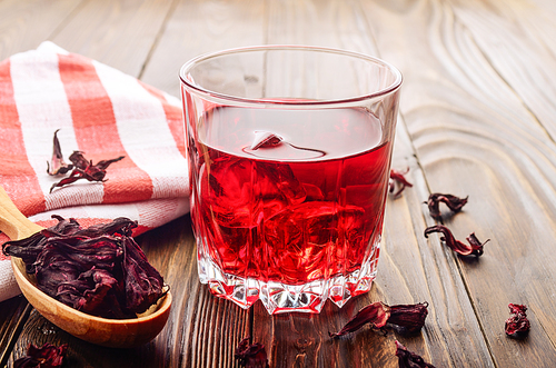 Closeup view at glass of tea with ice and spoon of dry hibiscus petals on wooden table background