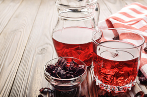 Closeup view at glass of hibiscus ice tea and jug on wooden table background