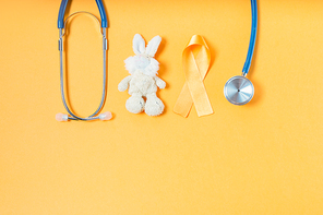 Children's toy rabbit with a Childhood Cancer Awareness yellow ribbon and stethoscope on yellow background with copy space, top view