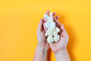Hands holding children's toy rabbit on yellow background with copy space