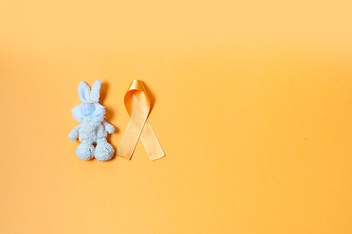 Children's toy rabbit with a Childhood Cancer Awareness yellow ribbon on yellow background with copy space, top view