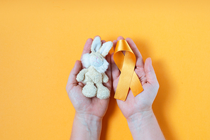 Hands holding children's toy rabbit with a Childhood Cancer Awareness yellow ribbon on yellow background with copy space. Childhood Cancer Day February, 15