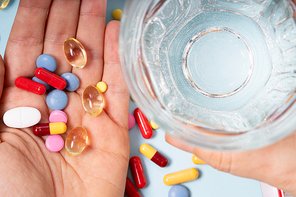 Someones hand holding colorful pills with glass of clear water over blue background. Medical pharmacy concept.