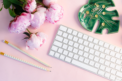 Flat lay top view home office workspace - modern keyboard with female accessories and peony flowers, copy space on pink background