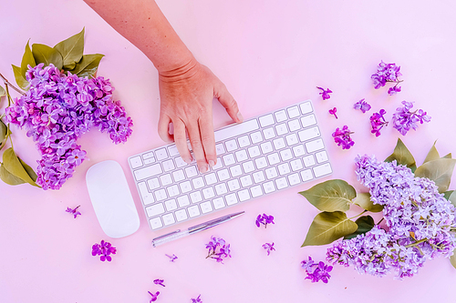 Flat lay home office workspace - modern keyboard with someone hand typing, fresh lilac flowers, toned