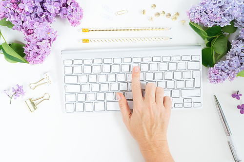 Flat lay top view home office workspace - hand typing on modern keyboard with lilac flowers on white background