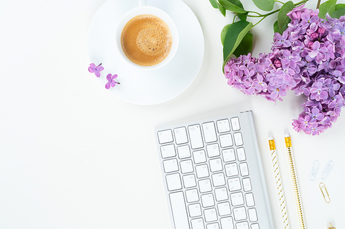Flat lay top view home office workspace - modern keyboard with cup of coffee and lilac flowers on white background
