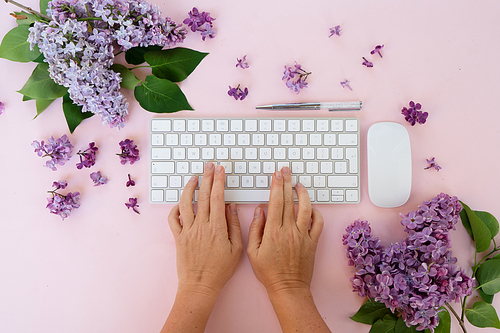 Flat lay home office workspace - modern keyboard with two hands typing, fresh lilac flowers