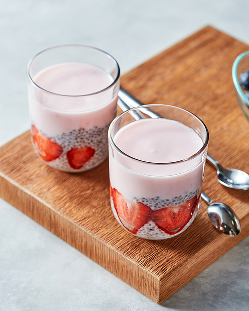 two glasses with smoothie and pieces of srtawberries, chia seeds on a wooden board on a gray background. concept of healthy ing food.