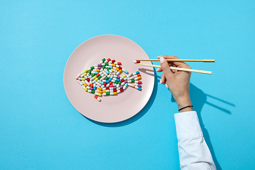Different pills and supplements as a fish on a white plate chopsticks in woman's hands with shadows on a blue. Suppressants for dieting concept. Top view.