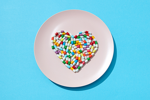Many different pills and supplements in a shape of a heart on a white plate on a blue background. Cardiovascular disease prevention. Flat lay
