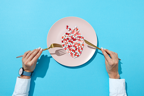 Fish from colorful pills and tablets on a white plate, girl's hands with watch on a blue background with shadows, copy space. The negative effect of pills on cardiovascular diseases. Top view.