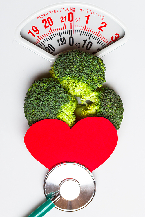Diet healthy eating weight control and health care concept. Closeup green broccoli with stethoscope and red heart on white scales