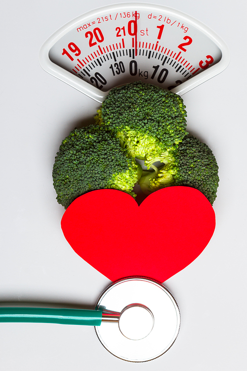 Diet healthy eating weight control and health care concept. Closeup green broccoli with stethoscope and red heart on white scales