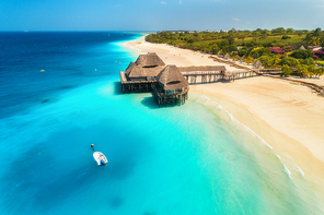 Aerial view of beautiful hotel on the water in ocean at sunset in summer. Zanzibar, Africa. Top view. Seascape with wooden hotel on the sea, boat, azure water, sandy beach, green palm trees. Resort