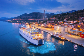 Aerial view of cruise ship at harbor at night. Landscape with ships and boats in harbour, city illumination, buildings, mountains, blue sea at sunset. Top view. Luxury cruise. Floating liner in port