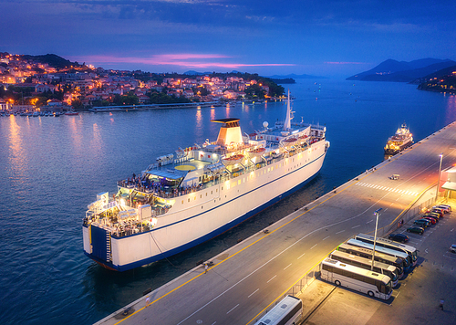 Aerial view of cruise ship at harbor at night. Landscape with ships and boats in harbour, city illumination, buildings, mountains, blue sea at sunset. Top view. Luxury cruise. Floating liner in port