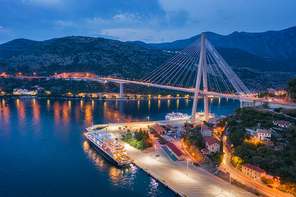Aerial view of cruise ship at harbor and beautiful bridge at night. Landscape with ships and boats in harbour, city illumination, road, mountains, blue sea at sunset. Top view. Floating liner in port