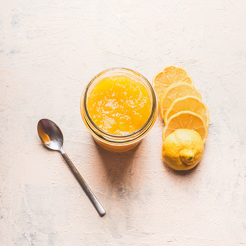 Lemon curd in glass with sliced lemon and spoon on white table, top view