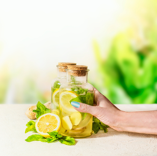 Female women hand holding bottle with ginger, lemon, mint lemonade on white table at green nature background. Healthy summer drinks. Natural Immun booster. Infused fruits water.
