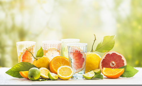 Various healthy summer lemonade in glasses with variety of organic citrus fruits: lime, oranges, grapefruit and lemon on table at green sunny garden nature background. Infused water. Copy space