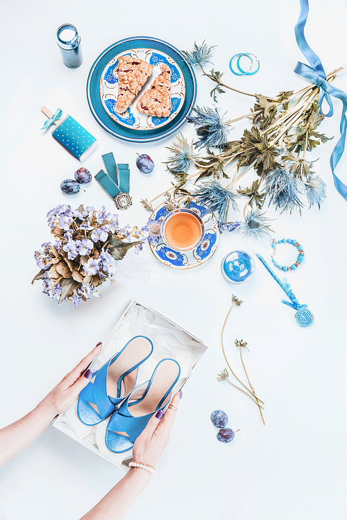 Woman hands holding new blue shoes in box on white desktop. Feminine still life  with cosmetic products, flowers bunch, cup of tea with cake pieces, jewelry. Top view. Flat lay. Fashion blogging