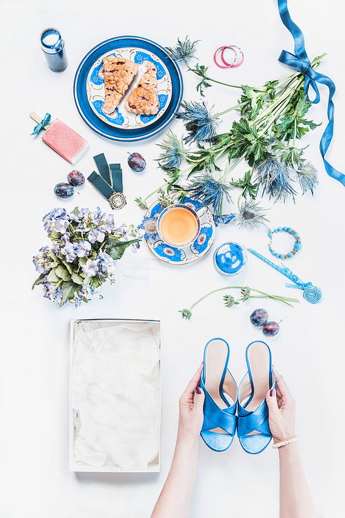 Woman hands holding new blue sandals on white desktop with empty box and feminine still life of cosmetic products, flowers bunch, cup of tea with cakes, jewelry.  Top view. Flat lay. Fashion blogging