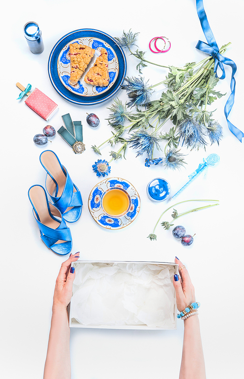 Woman hands holding empty box on white desktop with new blue sandals and feminine still life of cosmetic products, flowers bunch, cup of tea with cakes, jewelry.  Top view. Flat lay. Fashion blogging