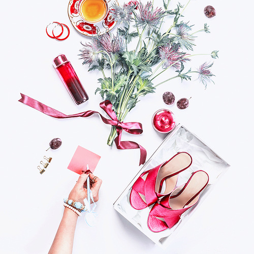 Woman hand writes a note on blank pink paper on white desk with high heels in box, cosmetics, perfume, flowers bunch, cakes cup of tea and bijou on white desk background. Casual woman beauty blog