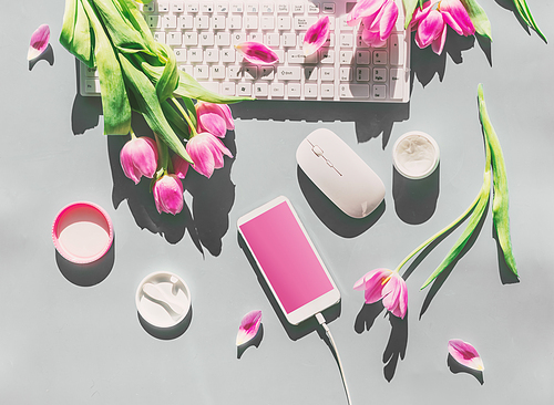 Female desktop with light pink tulips flowers bunch, cosmetics,  PC keyboard , smartphone with blank screen and wire . Top view. Mothers day . Springtime. Greeting