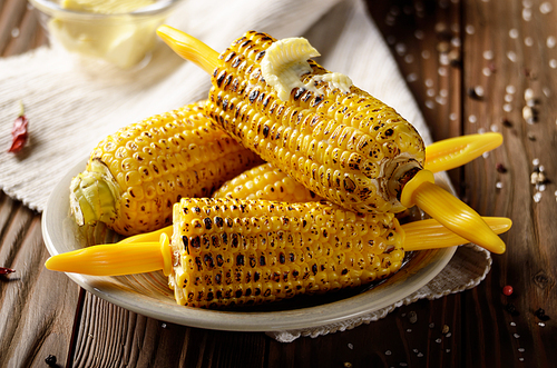 Wooden table with deep grilled sweet corn cobs under melting butter with plastic holder on clay dish