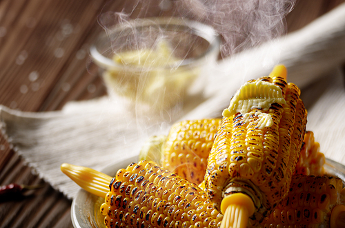 Wooden table with deep grilled sweet corn cobs under melting butter with plastic holder on clay dish