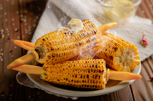 Wooden table with deep grilled sweet corn cobs under melting butter with plastic holder on clay dish