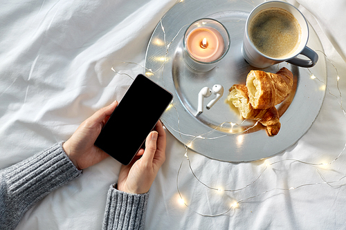 technology, hygge and christmas concept - hands of woman with smartphone, croissant and coffee in bed