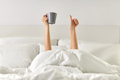 morning, comfort and people concept - young woman with cup of coffee lying in bed and showing thumbs up at home bedroom