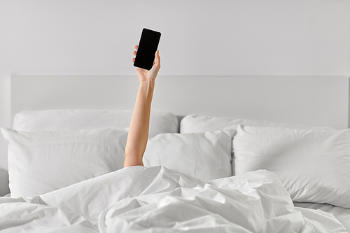 technology, comfort and morning concept - hand of young woman lying in bed with smartphone at bedroom