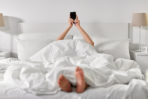 technology, comfort and morning concept - hands of young woman lying in bed with smartphone at bedroom