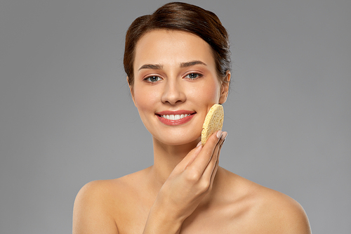 beauty, people and skincare concept - young woman cleaning face with exfoliating sponge over grey background