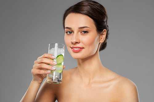 beauty and detox concept - woman drinking fresh water with cucumber and ice over grey background