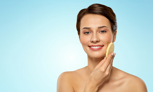 beauty, people and skincare concept - young woman cleaning face with exfoliating sponge over blue background