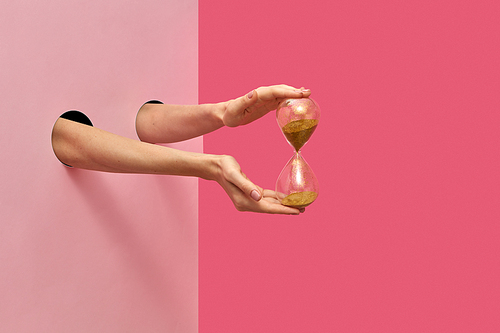 Vintage sandglass in a female's hands from two holes in the wall on a duotone pink background, copy space.