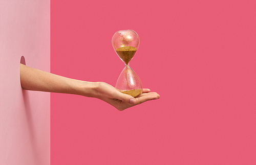 Woman's hand holds old hourglass in her hand through the hole in the wall on a duotone pink background with soft shadows, copy space.