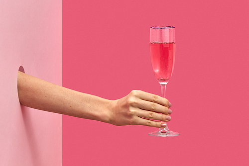 Holiday glass of rose wine in a female's hand through the hole in the wall on a duotone pink background with soft shadows, copy space. Holiday concept.