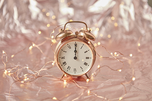 Golden alarm clock with time is midnight on a shiny cooper abstract background with string of lights, copy space. Greeting card.